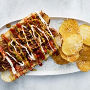 Top view of a loaded hot dog with various toppings served next to crispy potato chips on a plate.