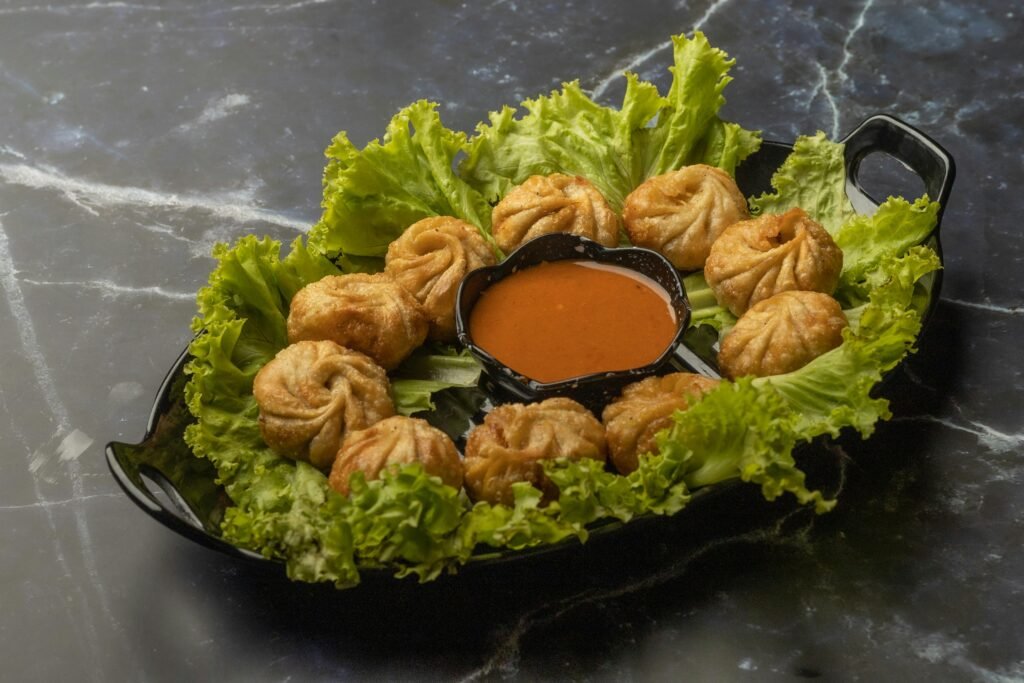 Crispy fried veg momos arranged with spicy sauce on a black platter, garnished with fresh lettuce.