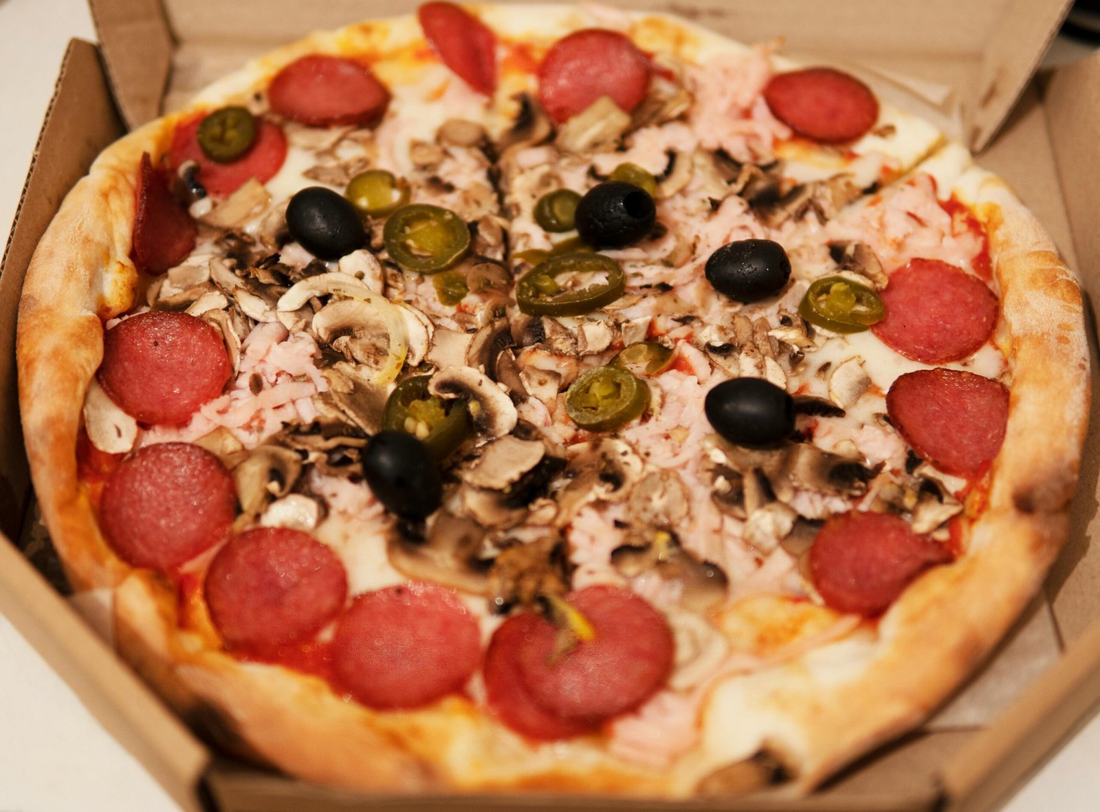 Close-up of a freshly baked pizza topped with pepperoni, mushrooms, olives, and jalapenos in a cardboard box.