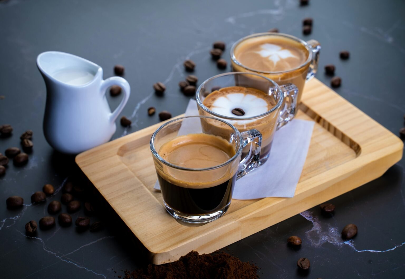 Three glasses of coffee with different toppings on a tray, accompanied by a milk creamer, creating a cozy beverage scene.